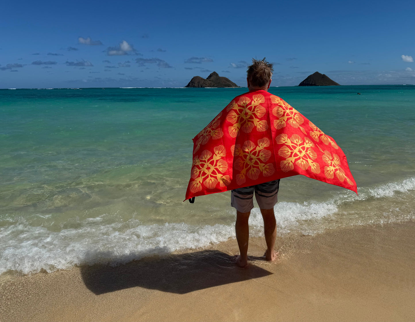 Person holding a red and gold patterned towel on a beach with turquoise water and distant islands.