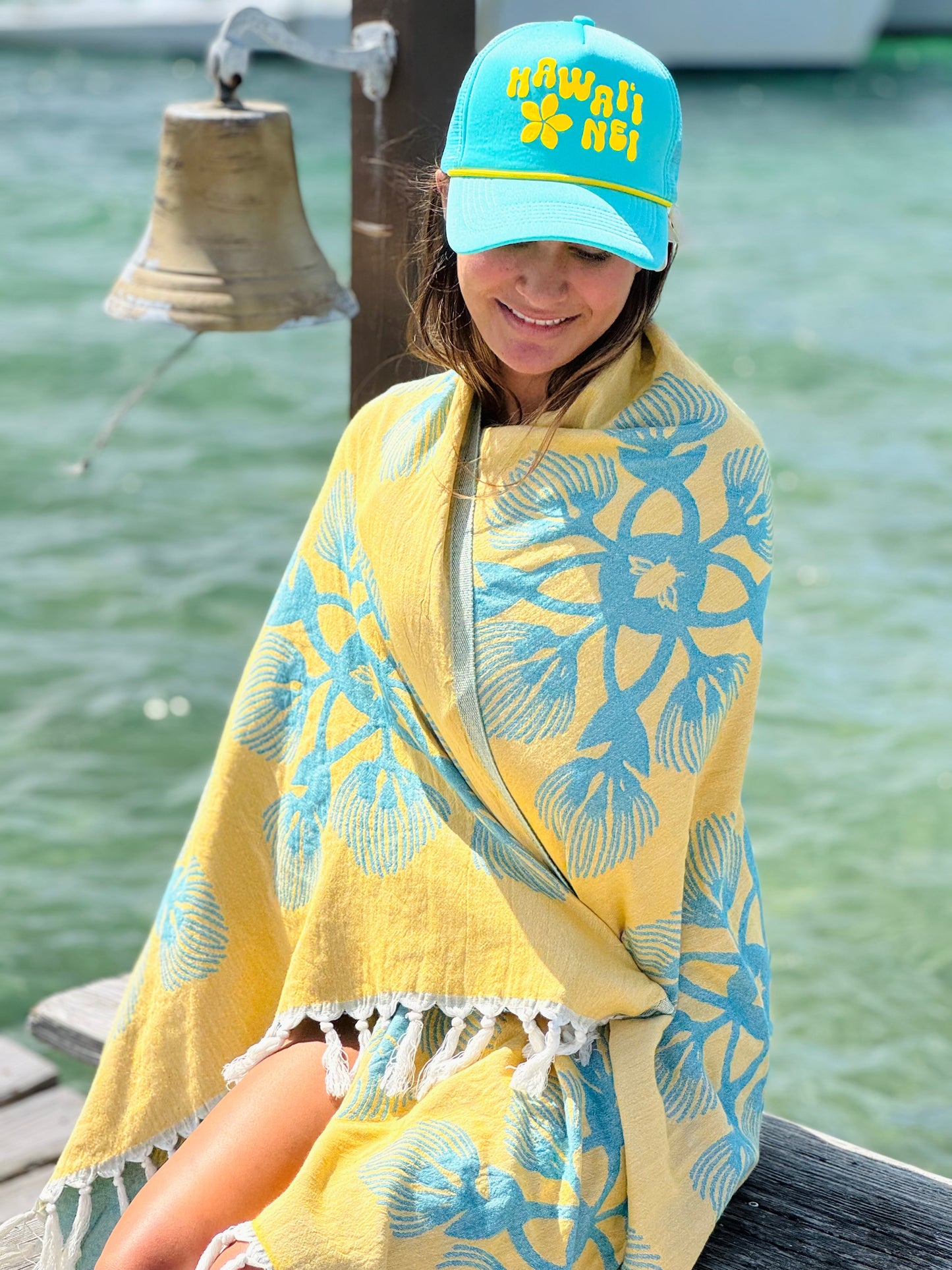 Person wrapped in a yellow and blue patterned towel wearing a matching cap by a bell and water.